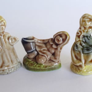 May include: Three small ceramic figurines. The first is a person holding a candle, the second is a reclining lamb with a bucket, and the third is a woman with a staff. All are in shades of tan, brown, and green.