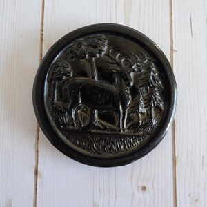 May include: A round, black decorative plate with a raised relief design. The relief depicts a deer standing in a forest scene with trees and foliage. The plate has a glossy finish and is set against a light wood background.