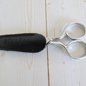 May include: A pair of silver scissors with a black leather sheath. The sheath has the word "gimphee" embossed on it. The scissors are on a light wood surface.