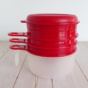 May include: Stack of three red measuring cups with handles and lids, resting on a clear, frosted bowl. The handles are designed for easy gripping. The set is designed for kitchen use.