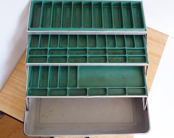 Puede incluir: Una caja de aparejos de metal vintage abierta, que revela tres bandejas de plástico verde y un gran compartimento inferior. Las bandejas están divididas en secciones rectangulares. La caja es plateada con un asa de metal. Ancho aproximado: 30 cm.