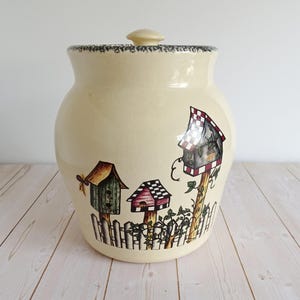 May include: A cream-colored ceramic canister with a matching lid, featuring a hand-painted scene of three colorful birdhouses on wooden posts behind a white picket fence, accented with green vines and a small butterfly on the left side.