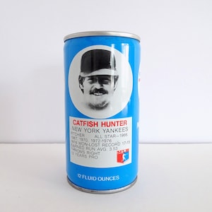 May include: A vintage blue can featuring a portrait of Catfish Hunter, a pitcher for the New York Yankees. The can includes text detailing his career stats, including his All-Star year, won-lost record, and earned run average. The can holds 12 fluid ounces.