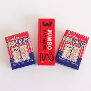 May include: Three rectangular cardboard boxes of fireworks. Two identical boxes are labeled Leader Brand Black Python Snakes, Made in Japan, Six Pcs No. 6. The center box is red and labeled 3 Jumbo Hat Snakes with a black snake illustration.