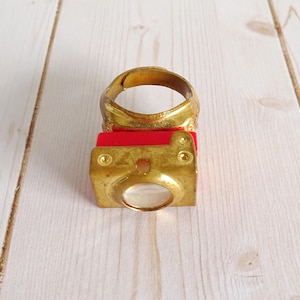 May include: A gold-colored ring with a camera design. The ring features a red rectangular base and a gold-colored camera body with a clear lens. The ring is set on a light wood surface.