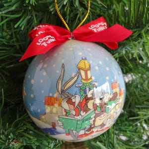 May include: A round Christmas ornament featuring Bugs Bunny and other Looney Tunes characters in a winter scene. The ornament has a red ribbon with "Looney Tunes" printed on it. The ornament is hanging on a Christmas tree.