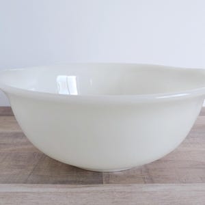 May include: Off-white mixing bowl with a rounded shape and two small handles. The bowl is made of a smooth, opaque material and sits on a wooden surface. The bowl is designed for food preparation.
