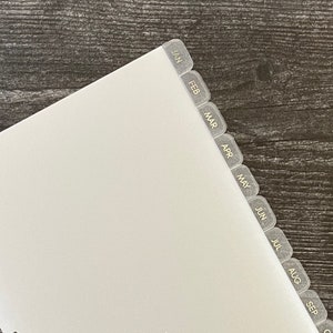 May include: A white planner insert with monthly tabs labeled January through December. The tabs are gray with black text. The insert has pre-punched holes for binding. The background is a dark wood grain.