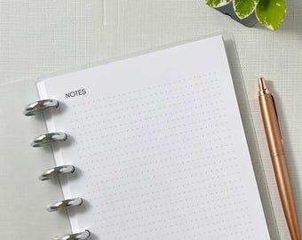 May include: A white notebook with dotted pages and the word "NOTES" printed at the top. A rose gold pen rests beside the open notebook. The notebook is held together with silver rings. A small potted plant is in the background.