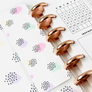 May include: Close-up of a planner with rose gold disc-bound rings. The planner pages have a pastel cloud pattern and a calendar grid. The planner is open, showing the month's calendar and the word "INSPIRAT".