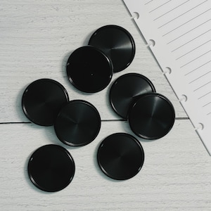 May include: A collection of eight black disc-shaped planner discs. The discs have a matte finish and a radial pattern on the surface. A white, lined paper with punched holes is visible in the background.