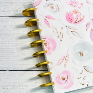 May include: A planner with a floral design featuring pink and gray watercolor flowers and gold accents. The planner is held together with gold disc-bound rings. The background is a white wooden surface.