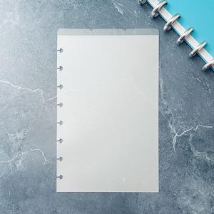 May include: A clear, frosted plastic planner divider with three labeled tabs at the top and pre-punched holes along the left edge. The divider is rectangular and sits on a gray marbled surface, with a blue planner binding visible in the corner.