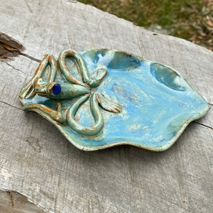 May include: A blue and green ceramic dish with a frog design. The frog has a blue glass eye.