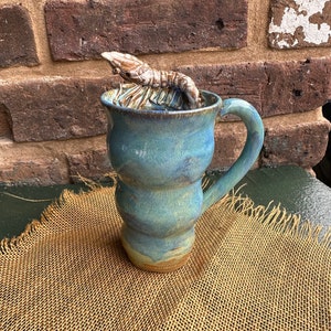 May include: A blue ceramic mug with a wavy design and a handle. A ceramic shrimp sits on top of the mug.