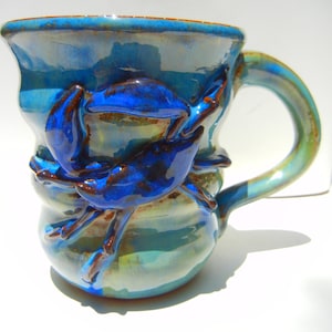 May include: A handmade ceramic mug with a blue glaze and a crab design. The mug features a curved handle and a detailed crab sculpture on the side. The colors include shades of blue, brown, and green.