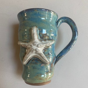 May include: A blue and green ceramic mug with a white starfish design. The mug has a handle and a glazed finish.