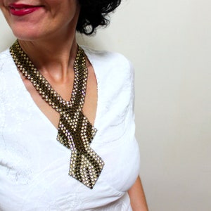 May include: A beaded necklace in a tie-like design. The necklace features a pattern of black, gold, and white beads. The necklace is worn over a white blouse.