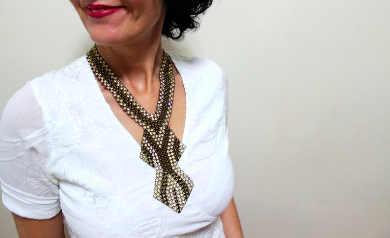 May include: A beaded necklace in a tie-like design. The necklace features a pattern of black, gold, and white beads. The necklace is worn over a white blouse.