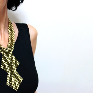 May include: A beaded necklace with a geometric design. The necklace features a pattern of alternating gold and black beads, creating a striking visual contrast. The necklace is worn against a black top, highlighting its intricate details.