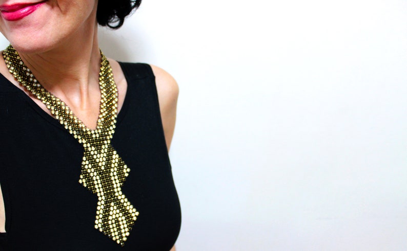 May include: A beaded necklace with a geometric design. The necklace features a pattern of alternating gold and black beads, creating a striking visual contrast. The necklace is worn against a black top, highlighting its intricate details.