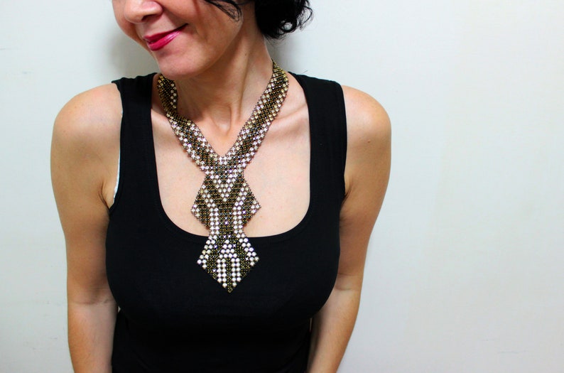 May include: A beaded necklace with a geometric design. The necklace features a combination of gold, white, and black beads, creating a striking pattern. The necklace is worn over a black tank top.