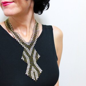 May include: A beaded necklace with a geometric design. The necklace features a combination of black, gold, and white beads, creating a striking contrast. The necklace is worn over a black top, and the background is plain white.