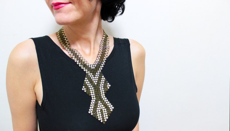May include: A beaded necklace with a geometric design. The necklace features a combination of black, gold, and white beads, creating a striking contrast. The necklace is worn over a black top, and the background is plain white.