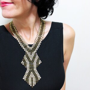 May include: A beaded necklace with a geometric design. The necklace features a combination of gold, black, and white beads, creating a striking contrast. The necklace is worn over a black top.