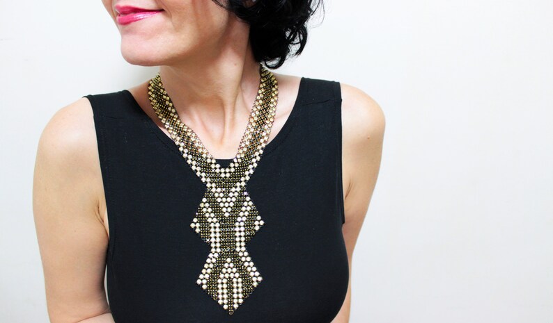 May include: A beaded necklace with a geometric design. The necklace features a combination of gold, black, and white beads, creating a striking contrast. The necklace is worn over a black top.