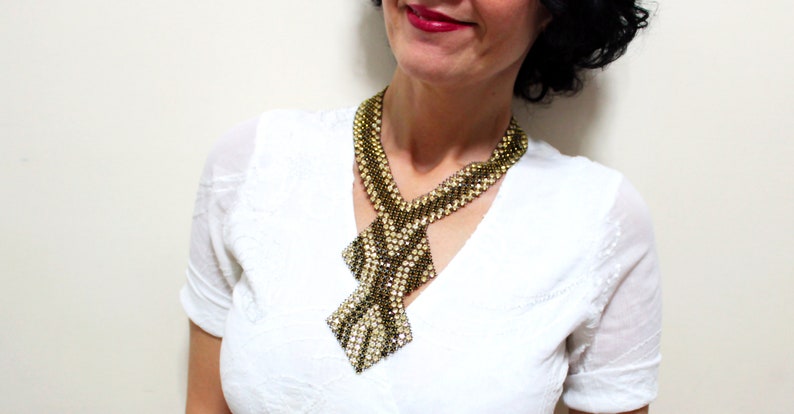 May include: A beaded necklace with a geometric design. The necklace features a combination of gold and silver beads, creating a textured pattern. The necklace has a bib-style design with a diamond-shaped pendant.