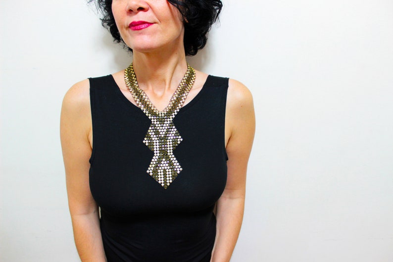 May include: A statement necklace with a geometric design. The necklace features a combination of gold, silver, and white beads, creating a striking contrast. The woman is wearing a black dress.