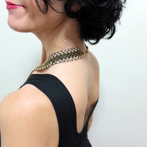 May include: A gold-toned necklace with clear rhinestones and a black backing is worn over a black top. The necklace is a wide band that sits around the neck. The woman is seen from the side, with her back to the camera.
