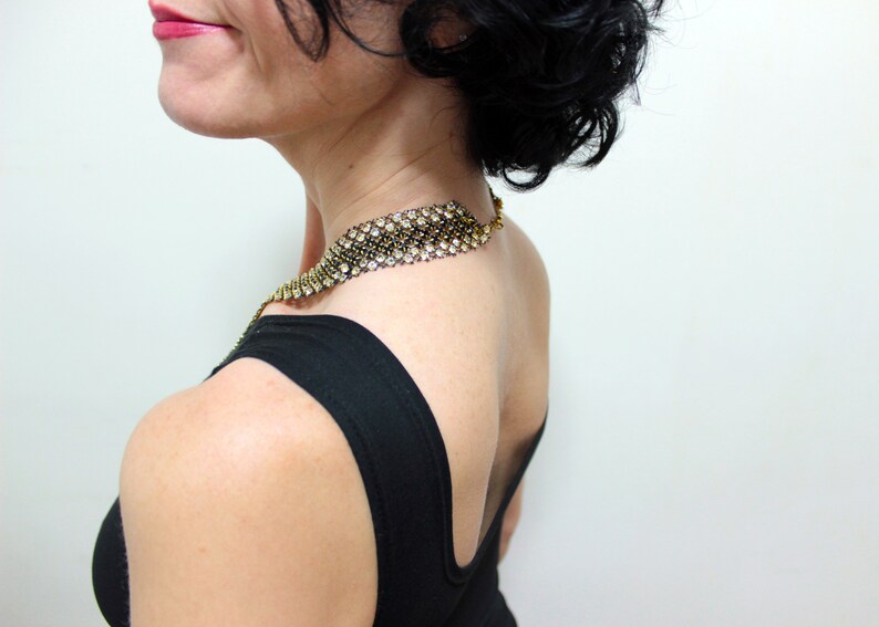 May include: A gold-toned necklace with clear rhinestones and a black backing is worn over a black top. The necklace is a wide band that sits around the neck. The woman is seen from the side, with her back to the camera.