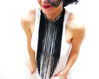 May include: A woman wearing a white tank top with a long black fringe necklace. The necklace has a silver chain at the top.