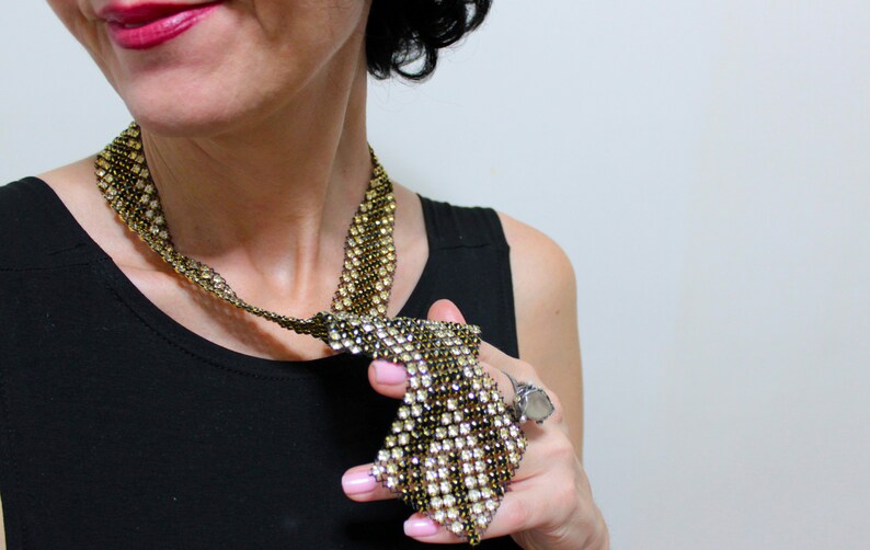 May include: A gold and clear rhinestone necklace with a unique, geometric design. The necklace is made of small, sparkling rhinestones set in a gold-tone metal. The necklace is worn over a black top.