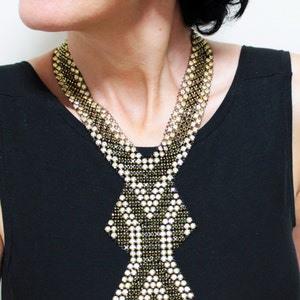 May include: A statement necklace featuring a geometric design. The necklace is composed of small, sparkling beads in gold, black, and white, creating a layered effect. The pendant has a diamond-like shape. The necklace is worn over a black top.