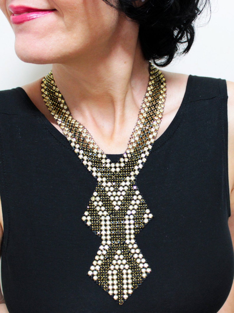 May include: A statement necklace featuring a geometric design. The necklace is composed of small, sparkling beads in gold, black, and white, creating a layered effect. The pendant has a diamond-like shape. The necklace is worn over a black top.