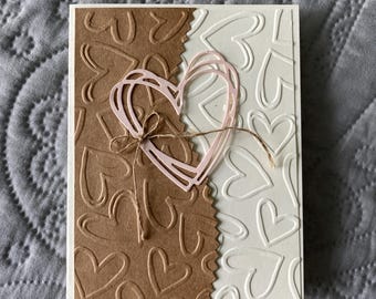 Könnte beinhalten: Eine handgemachte Karte mit braunem und weißem Hintergrund, die ein Herzmotiv zeigt. Das Herz ist aus rosa Papier und liegt auf dem Hintergrund. Die Karte ist mit einem braunen Bindfaden gebunden.