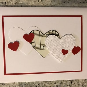 May include: A white card with a red border features a design of layered paper hearts. The hearts are decorated with red hearts and a piece of sheet music.