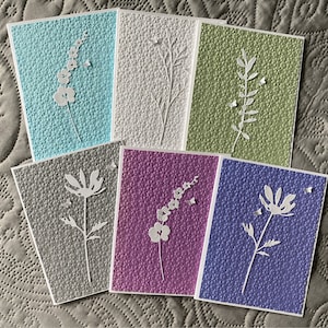 May include: A collection of handmade greeting cards with embossed floral designs. Each card features a different color, including blue, white, green, gray, purple, and lavender. The cards have a textured surface and white floral cutouts.