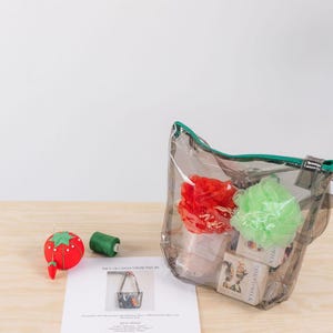 K&ouml;nnte beinhalten: Eine transparente Plastikt&uuml;te mit gr&uuml;nem Rei&szlig;verschluss und gr&uuml;nem Trageriemen. Die Tasche ist gef&uuml;llt mit Badesachen, darunter zwei Badeschw&auml;mme, einer rot und einer gr&uuml;n, und mehrere Seifenst&uuml;cke. Die Tasche steht auf einem Holztisch neben einem roten und gr&uuml;nen Nadelkissen und einer Spule gr&uuml;nen Fadens. Der Text "How to Sew a Striped Tote Bag" ist auf einem wei&szlig;en Blatt Papier gedruckt.