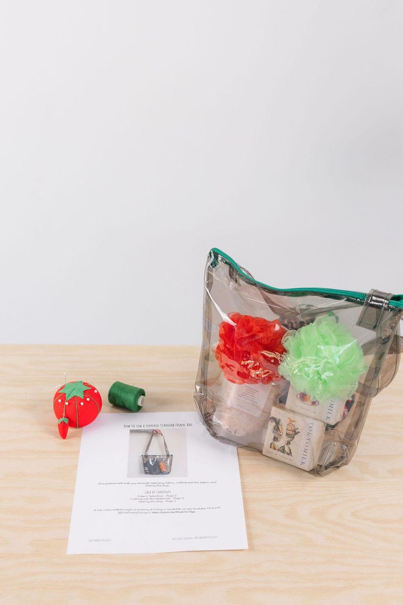 K&ouml;nnte beinhalten: Eine transparente Plastikt&uuml;te mit gr&uuml;nem Rei&szlig;verschluss und gr&uuml;nem Trageriemen. Die Tasche ist gef&uuml;llt mit Badesachen, darunter zwei Badeschw&auml;mme, einer rot und einer gr&uuml;n, und mehrere Seifenst&uuml;cke. Die Tasche steht auf einem Holztisch neben einem roten und gr&uuml;nen Nadelkissen und einer Spule gr&uuml;nen Fadens. Der Text "How to Sew a Striped Tote Bag" ist auf einem wei&szlig;en Blatt Papier gedruckt.