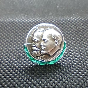 May include: A silver-toned pin featuring two profile portraits, possibly of historical figures, set against a globe design. The pin is encircled by a green laurel wreath, adding a touch of color and detail to the design.