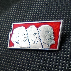 May include: A rectangular metal pin featuring a vibrant red enamel background. The design displays three silver-toned relief portraits of bearded figures facing right, arranged side-by-side within the silver-bordered frame of the pin.