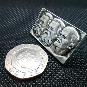 May include: A rectangular silver-toned metal pin featuring a relief sculpture of three bearded men in profile. Next to the pin is a silver-colored British twenty pence coin, which features the profile of Queen Elizabeth II and the text ELIZABETH II D G REG F D.