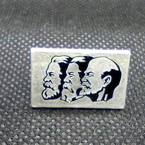 May include: A rectangular silver-toned metal pin featuring a black engraved graphic of three side-profile portraits of men with beards, arranged in a row. The design shows the distinct facial features and hairstyles of each individual figure.