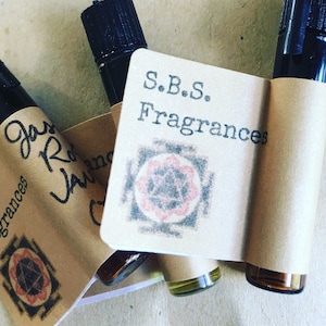 May include: Three small glass bottles containing brown liquid, each with a black cap. The bottles are labelled with a brown paper tag that reads "S.B.S. Fragrances" and features a pink and black mandala design.