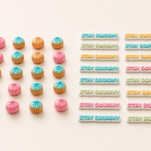 May include: A collection of small, round, bun-shaped charms in pink, orange, and blue, arranged in a grid alongside rectangular charms featuring the embossed text STAY SQUISHY in various colors including blue, yellow, pink, and green.