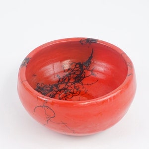 May include: A round, red ceramic bowl with a smooth finish. The interior and exterior are a solid red, with black, abstract line art designs on the inside. The bowl is set against a white background.